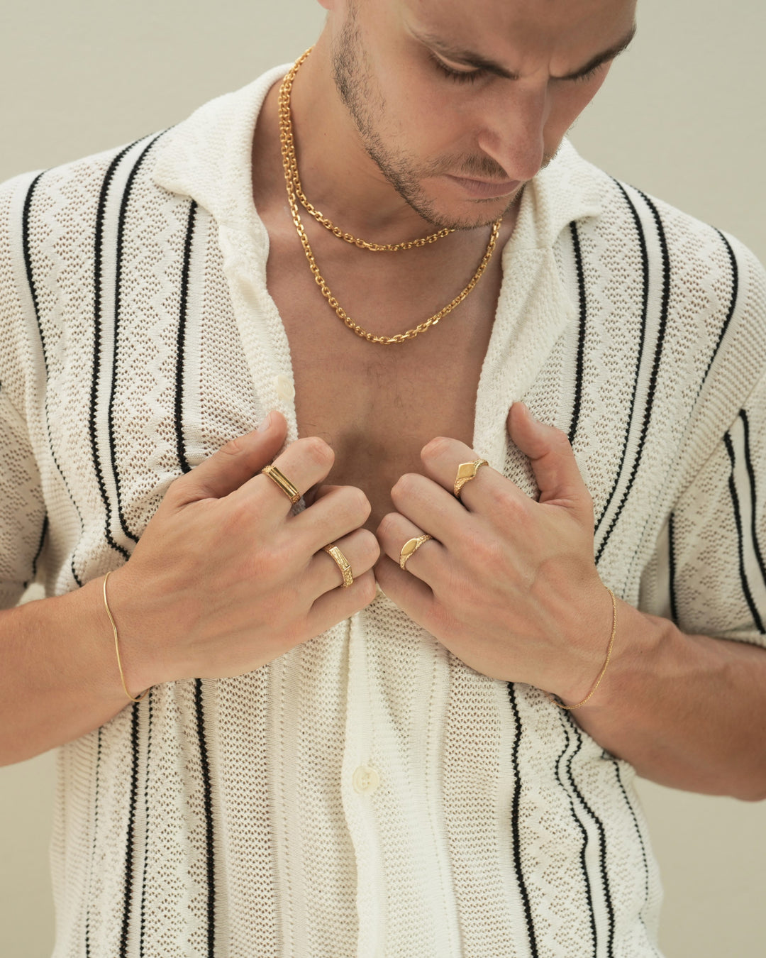 Founder of Nusands Corey Micari front view looking down to the left wearing 18k gold layered shackled necklaces, hands both grasping the inside of button up shirt at chest hieght. Right hand wearing roped in bracelet, line in the sand and six base rings, left hand wearing signature chain, suns out and mirrored signet rings.