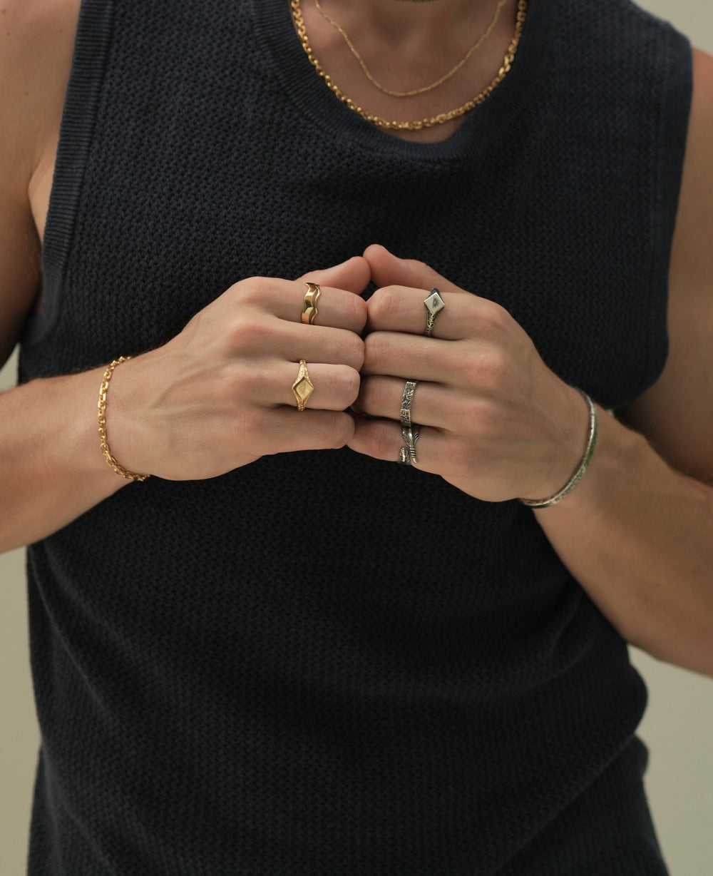 Founder of Nusands Corey Micari zoomed in chest shot wearing navy singlet, both hands meeting together in fists. Wearing 18K Gold stacked signature chain and shackled necklaces around neck, shackled bracelet on right wrist with suns out ring on a finger, stacked wave rings on another. His left arm is all sterling silver jewellery, stirking cuff on wrist, suns out, stacker and blade rings on hand.