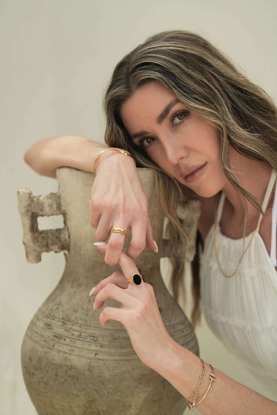 Founder of Nusands Emily Micari crouched over pot, head learning on right arm that is wearing 18K gold modest cuff and stacked wave rings, slightly holding left hand that is wearing midnight onyx ring on that finger.