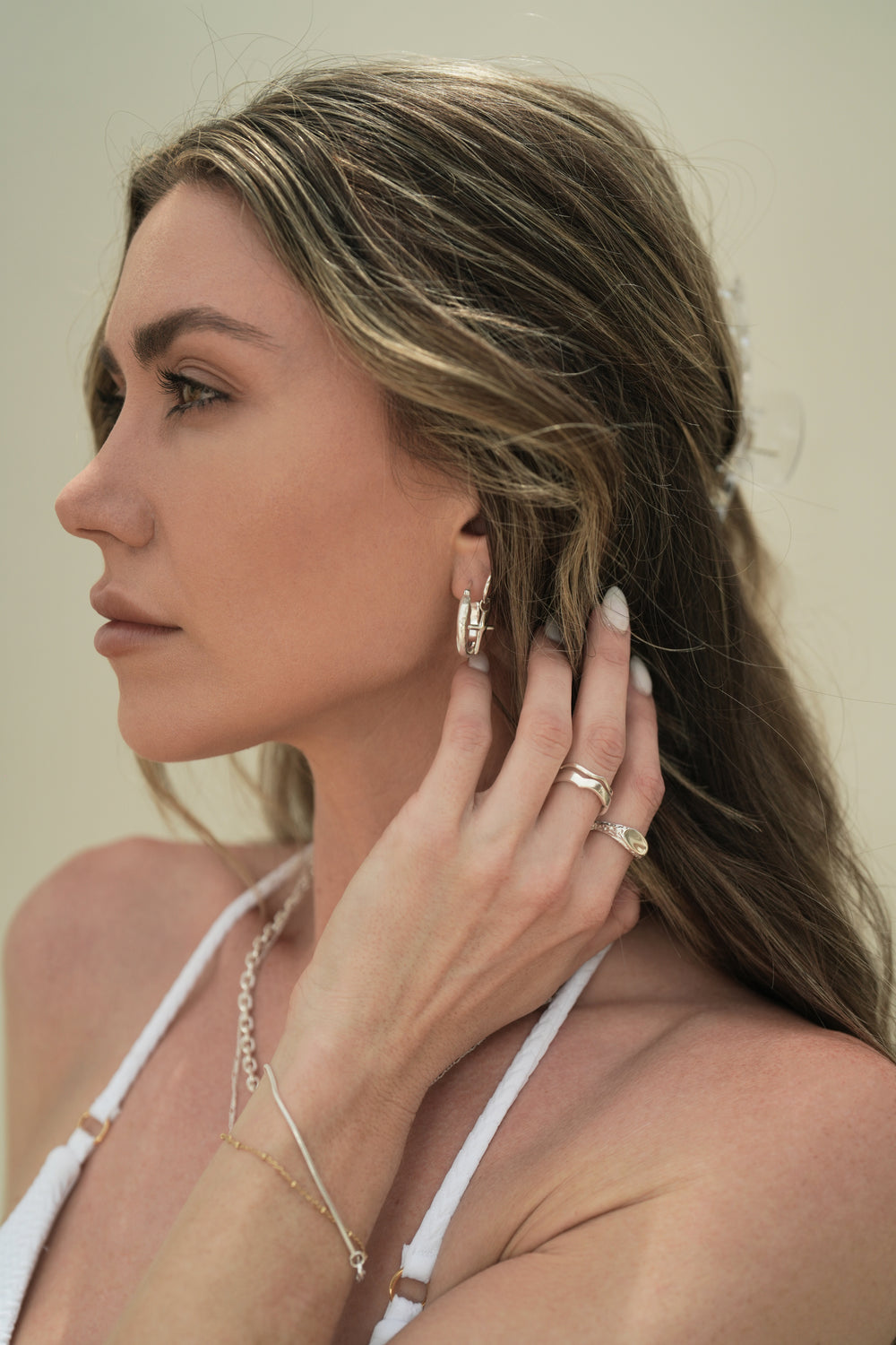 Founder of Nusands Emily Micari side profile of head wearing sterling silver stacked wavy and hoop earrings with cross charm in left eart. Left hand to side of neck showing stacked wave rings, mirrored signet ring, roped in bracelet and shackled necklace around her neck.