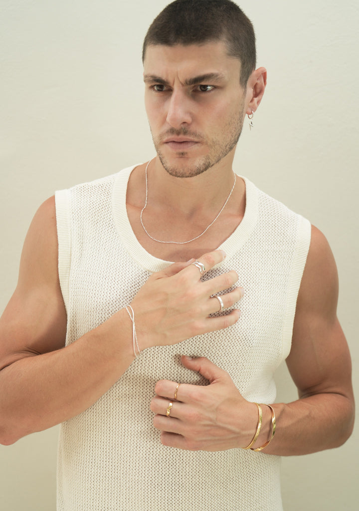 Founder of Nusands Corey Micari front on chest view looking slight to the right of camera. Sterling silver lightning bolt hoop earring in left ear, signature chain necklace around nck, stacked roped in and signature chain bracelets on right wrist, stacked wave rings on index finger and stacker ring on ring finger, hand rest on chest. Left wrist has stacked 18k gold modest and striking cuffs, unstacked wave rings on 2 fingers, hand resting lower than right hand on stomach