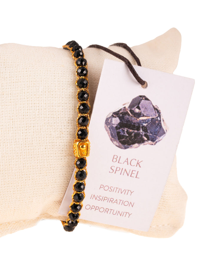 Black Spinel Gemstone Adjustable bracelet covering white cushion strung together with 18K Gold Yarn, Information tag showing.