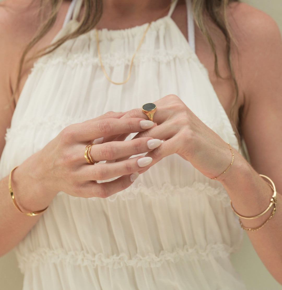 Founder of Nusands Emily Micari, zoomed in front on view of her chest, wearing a white dress with her hands slightly clasped together. Wearing 18k Gold jewellery, roped in necklace around her neck, Right hand has stacked wave rings around her middle finger, modest cuff on right wrist. Left hand has Midnight Onyx Ring on middle finger. Stacked gold jewellery around her left wrist.