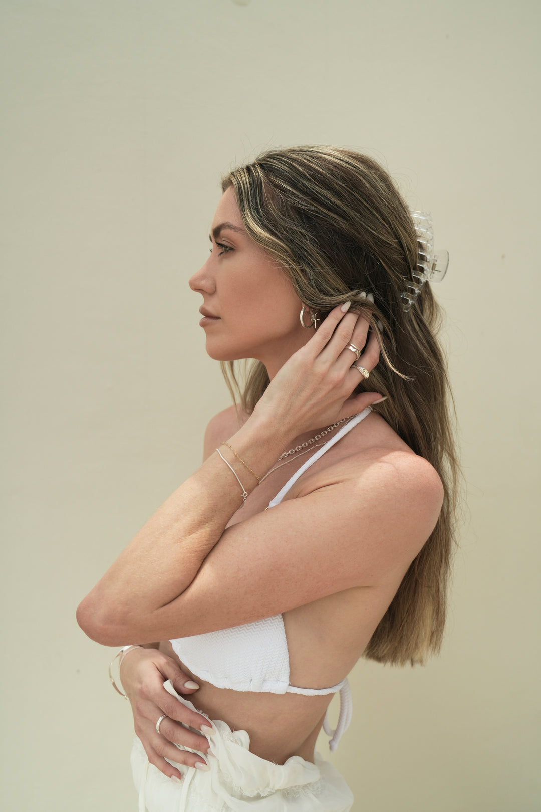 Founder of Nusands Emily Micari side profile left arm to back of neck, sterling silver stacked wave rings on middle finger, mirrored signet ring on index finger, shackled necklace around her neck, hoop earrings with cross charm in left ear, roped in bracelet on left wrist.