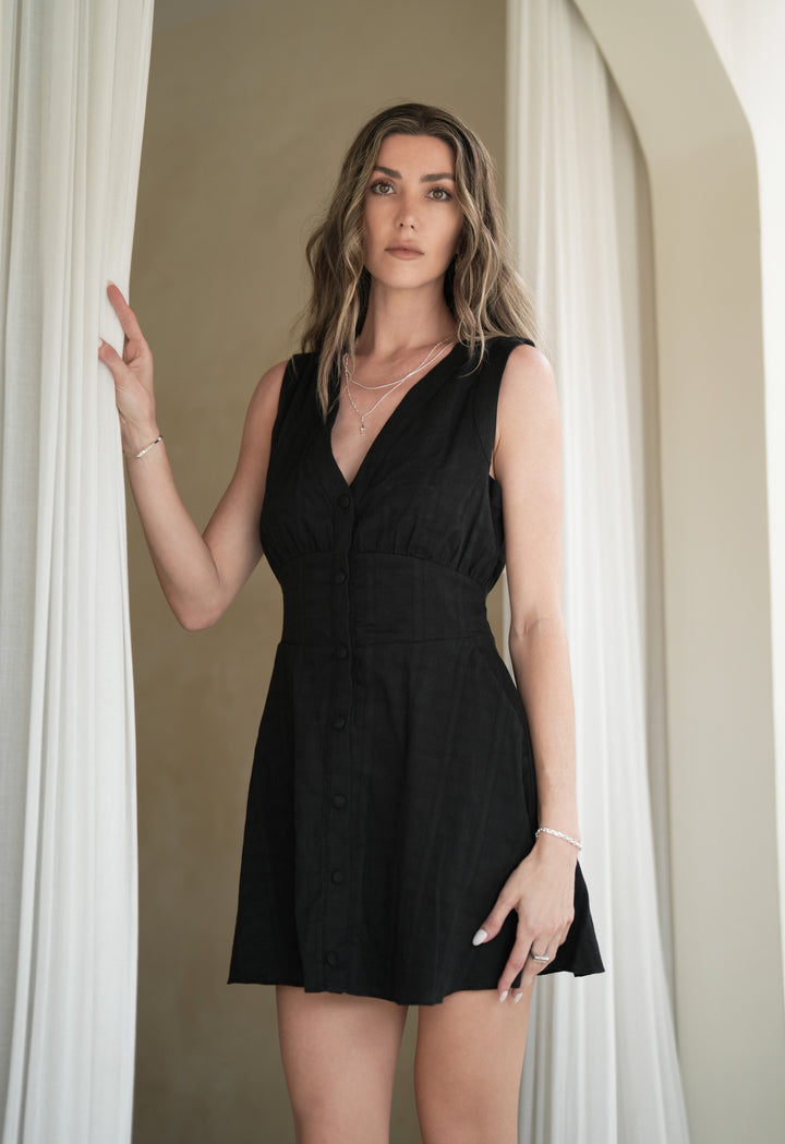 Founder of Nusands Emily Micari front profile standing in black dress left hand by her side wearing sterling silver shackled bracelet and line in the sand ring, right hand grasping curtain wearing modest cuff and wave rings, signature chain necklace with lightning bolt charm and roped in necklace around her neck, staring at camera.