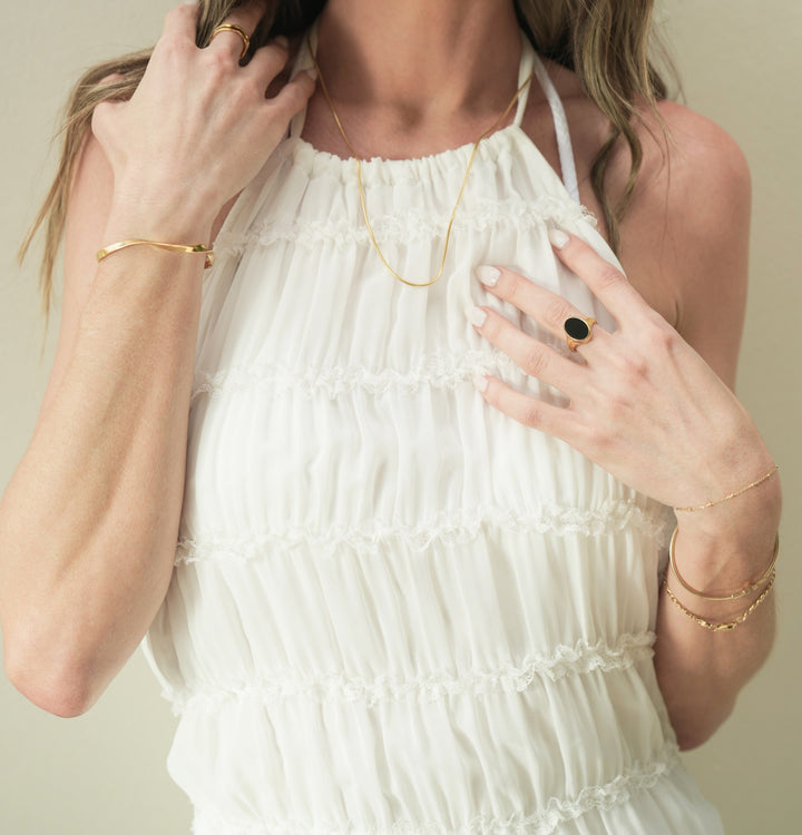 Founder of Nusands Emily Micari, zoomed in front on view chest shot, white dress. Wearing 18k Gold jewellery, roped in necklace around her neck, Right hand grabbing her right shoulder with stacked wave rings around her middle finger, modest cuff on right wrist. Left hand touching her chest, Midnight Onyx Ring on middle finger. Stacked gold jewellery around her left wrist.