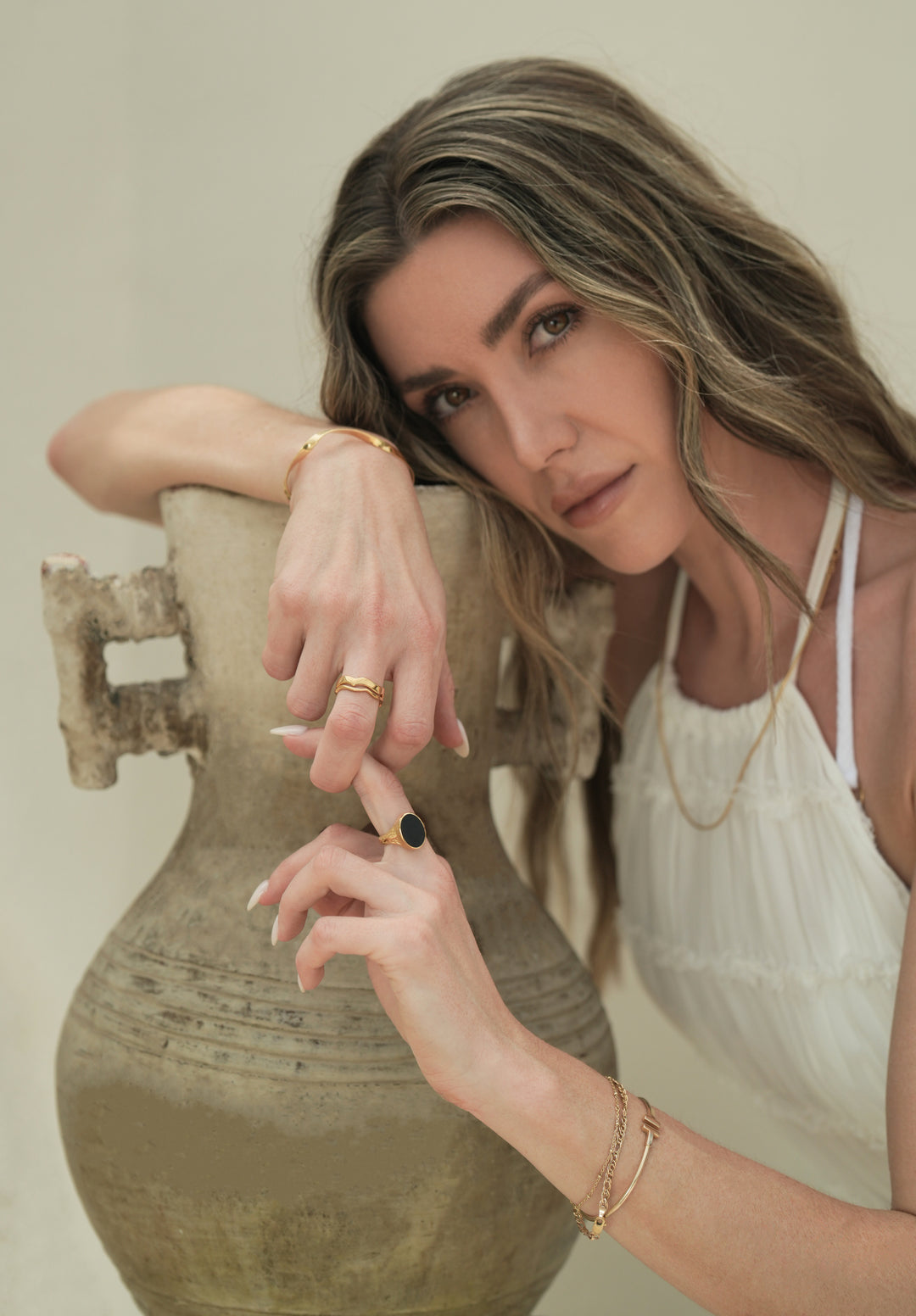 Founder of Nusands Emily Micari chest up view, looking at camera, right arm on a pot, head learning on right arm. Wearing 18k gold jewellery, modest cuff around right wrist, stacked wave rings on right middle finger, slightly grasping left hands middle finger that has Mindnight Onyx Ring on it. Stacked gold jewellery around left wrist, roped in necklace around her neck.