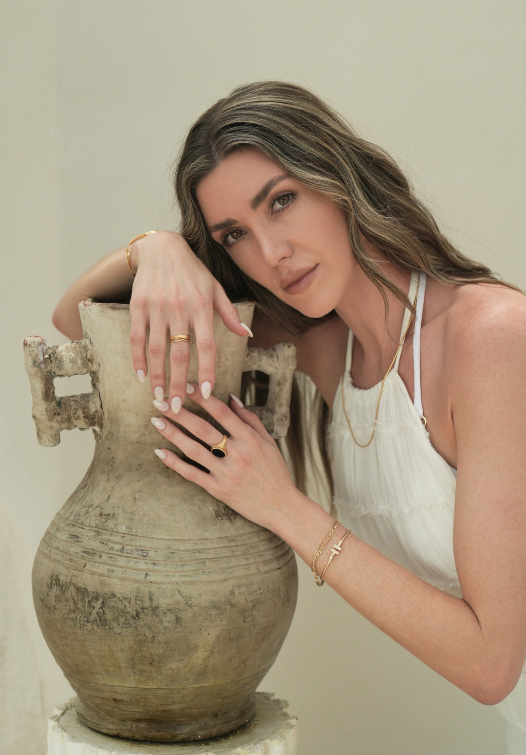 Founder of Nusands Emily Micari chest up view, looking at camera, right arm on a pot, head learning on right arm. Wearing 18k gold jewellery, modest cuff around right wrist, stacked wave rings on right middle finger, slightly grasping left hands middle finger that has Mindnight Onyx Ring on it. Stacked gold jewellery around left wrist, roped in necklace around her neck.