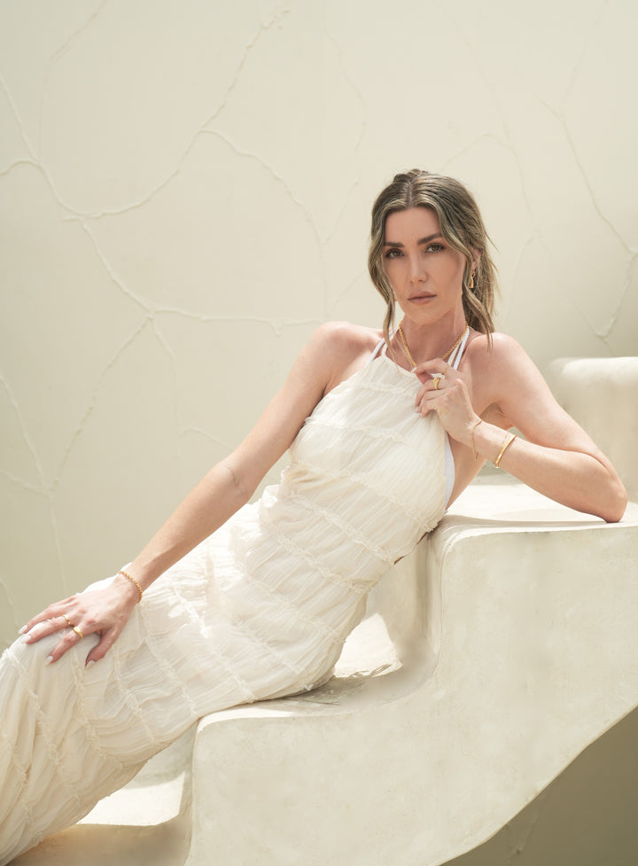 Founder of Nusands Emily Micari laying on stairs, white dress, full body, looking to camera, left arm holding 18k gold necklace that is around her neck, roped in bracelet around wrist, stacked with striking cuff. Right wrist by her side, shackled beacelet around it.