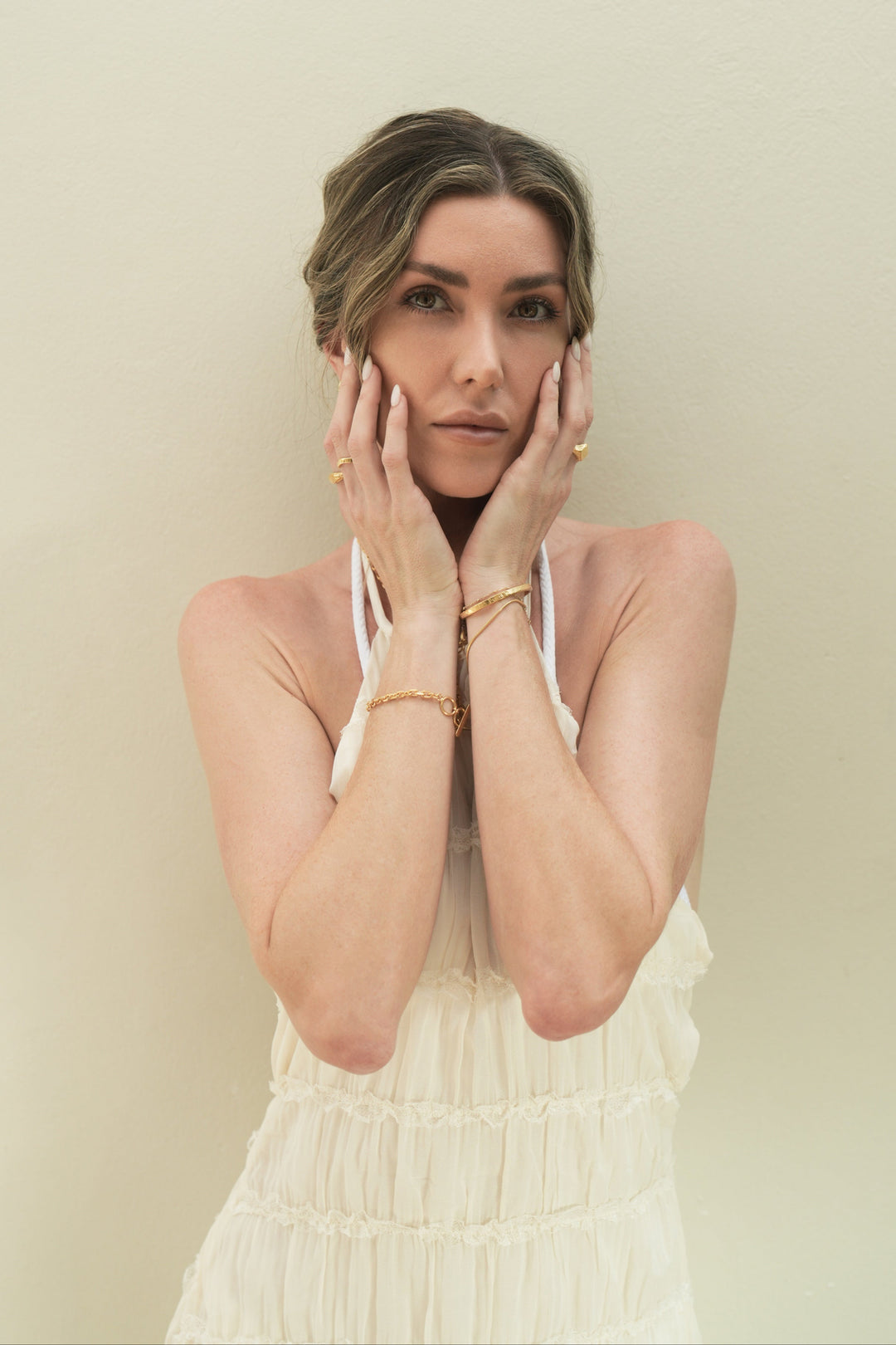 Founder of Nusands Emily Micari front profile both hand on the sides of her face, looking to camera. Left arm wearing 18k gold striking cuff stacked with roped in bracelet, and suns out ring. Right arm wearing shackled bracelet and wave rings.