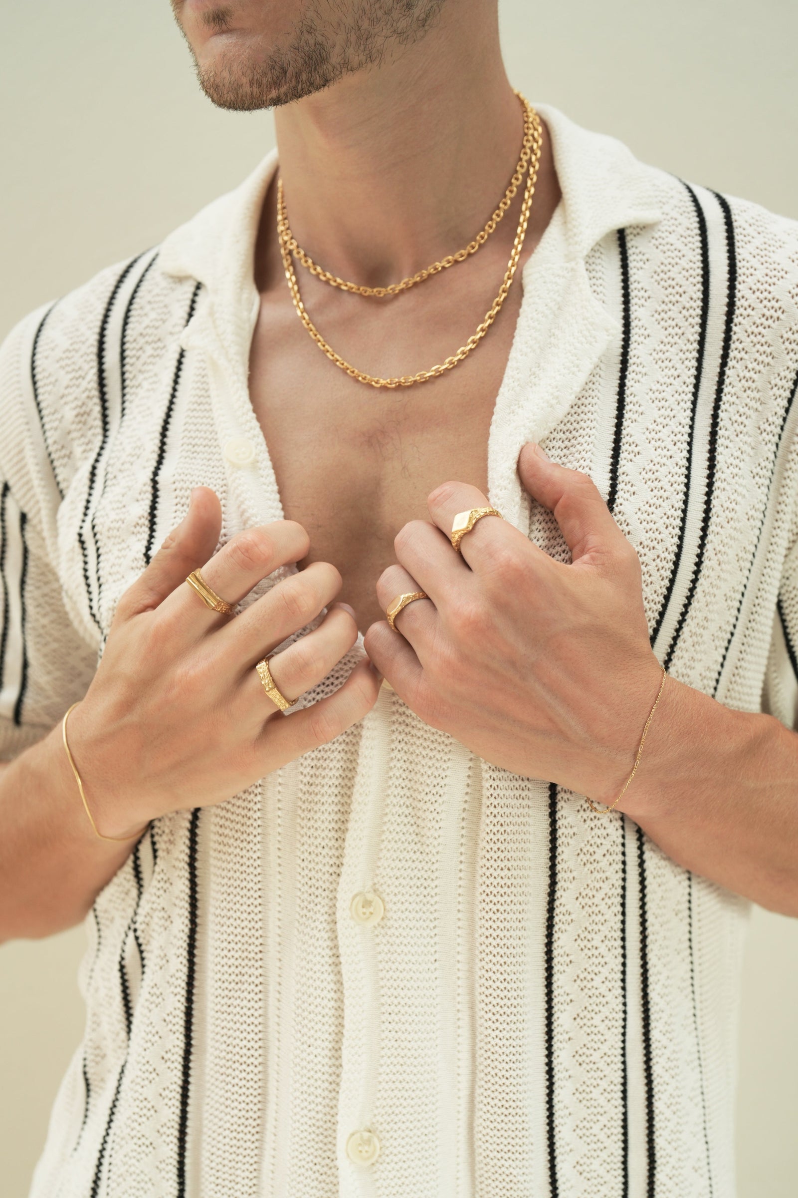 Founder of Nusands Corey Micari front chest view, hands grasping inside of half unbuttoned white and black striped shirt. Stacked 45 and 55cm 18k gold shackled necklaces around his neck. Signature chain bracelet on left wrist, suns out and mirrored signet on separate fingers. Right wrist has roped in bracelet on with line in the sand and six base rings on separate fingers.