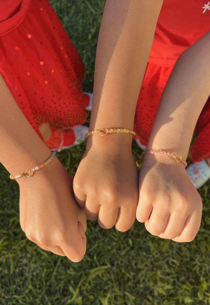 Three kids hands wearing rainbow, gold and pink gemstone bracelets with a grassy background