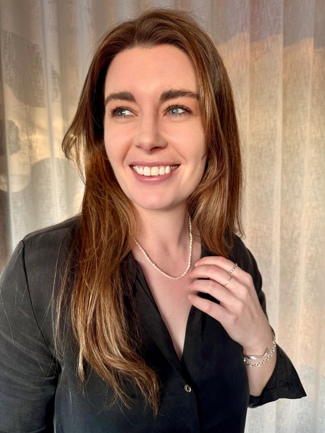 Woman with long brown hair wearing Nusands Pearl necklace and other silver jewellery in a black top against a neutral background