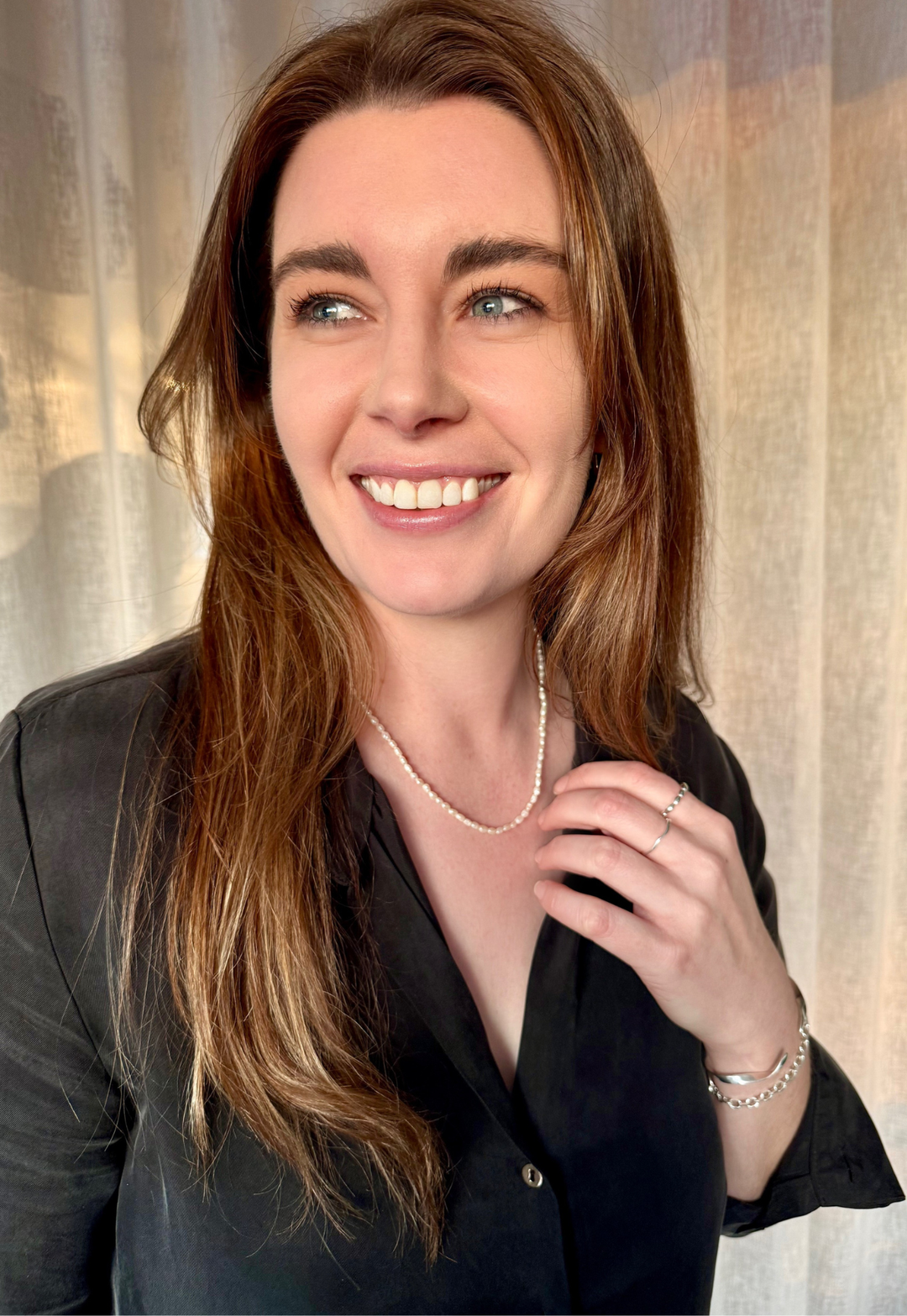 Woman with long brown hair wearing Nusands Pearl necklace and other silver jewellery in a black top against a neutral background