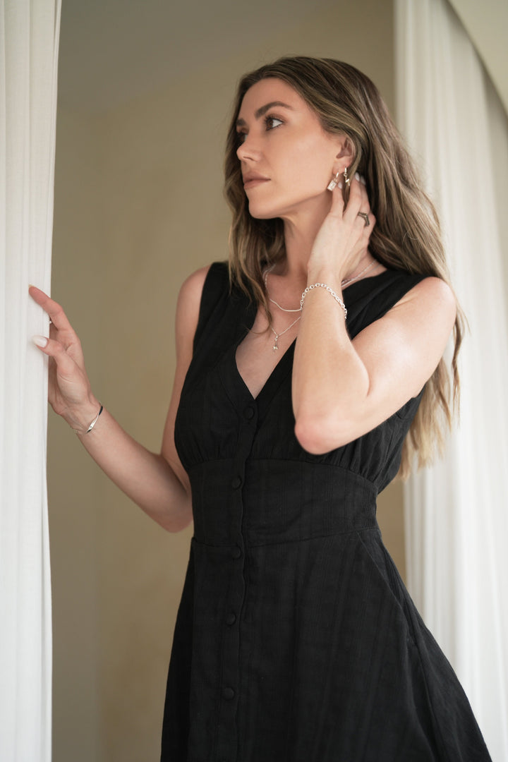 Founder of Nusands Emily Micari waist up front on view of her wearing a black dress, looking to the right of camera. Right arm grasping a curtain sterling silver modest cuff on her wrist, left arm lightly grasping the back of her neck, shackled bracelet on her wrist, line in the sand ring on her finger, stacked evil eye and lightning bolt earrings in her left ear. Roped in necklace stacked with signature chain necklace and lightning bolt charm. 