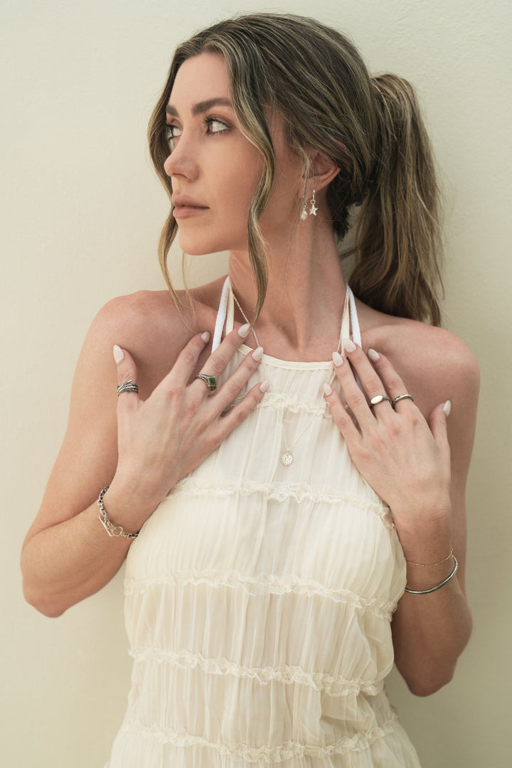 Founder of Nusands Emily Micari front view looking over to the right. wearing stacked sterling silver pearl and star earrings in her left ear. Both hands open to her chest striking cuff and signature chain bracelet stacked on left wrist, mirrored signet and stacker on left hand. Right wrist with shackled bracelet with blade and suns out ring with signature chain necklace and moon charm around her neck.