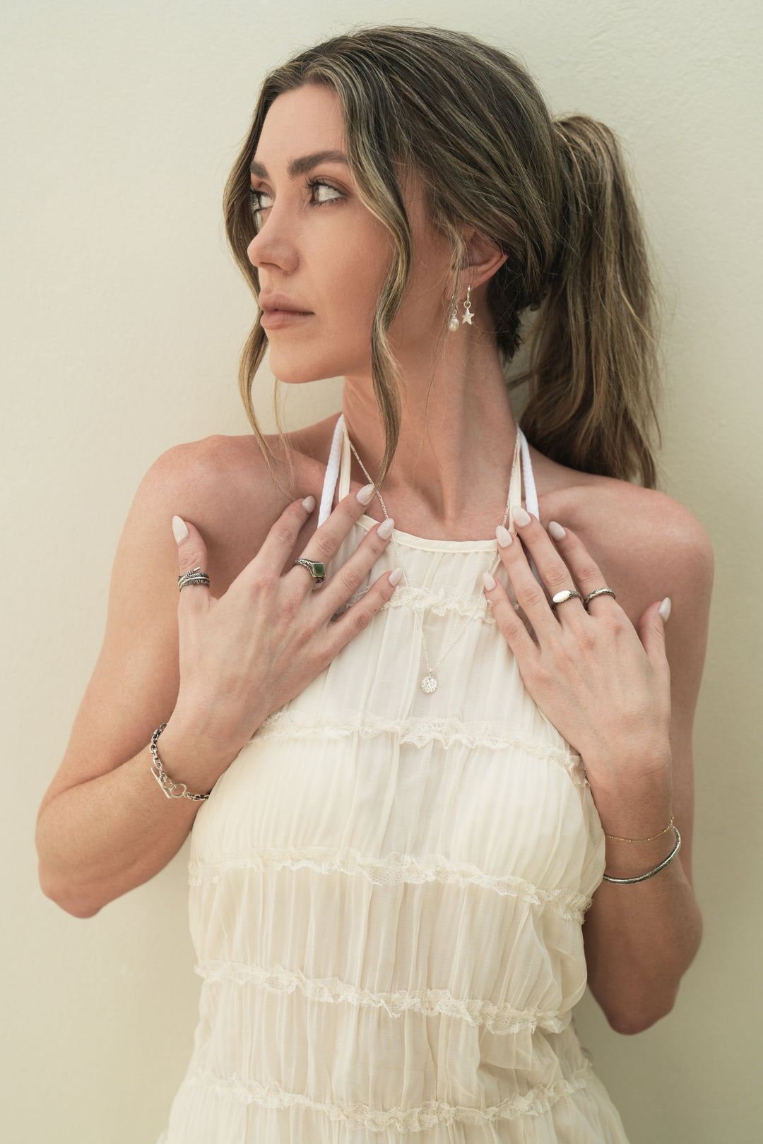 Founder of Nusands Emily Micari front view looking over to the right. wearing stacked sterling silver pearl and star earrings in her left ear. Both hands open to her chest striking cuff and signature chain bracelet stacked on left wrist, mirrored signet and stacker on left hand. Right wrist with shackled bracelet with blade and suns out ring with signature chain necklace and moon charm around her neck.