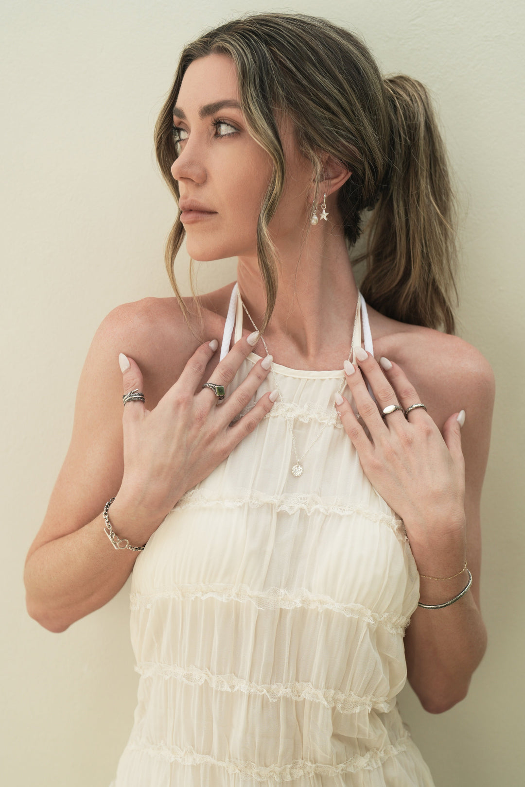 Founder of Nusands Emily Micari, looking away hands both up to chest wearing Sterling Silver earrings pearl and star charms in left ear, signature chain necklace with moon charm around neck, striking cuff and signature chain bracelet with mirrored signet and stacker ring on left arm, shackled bracelet, blade and suns out rings on right hand.