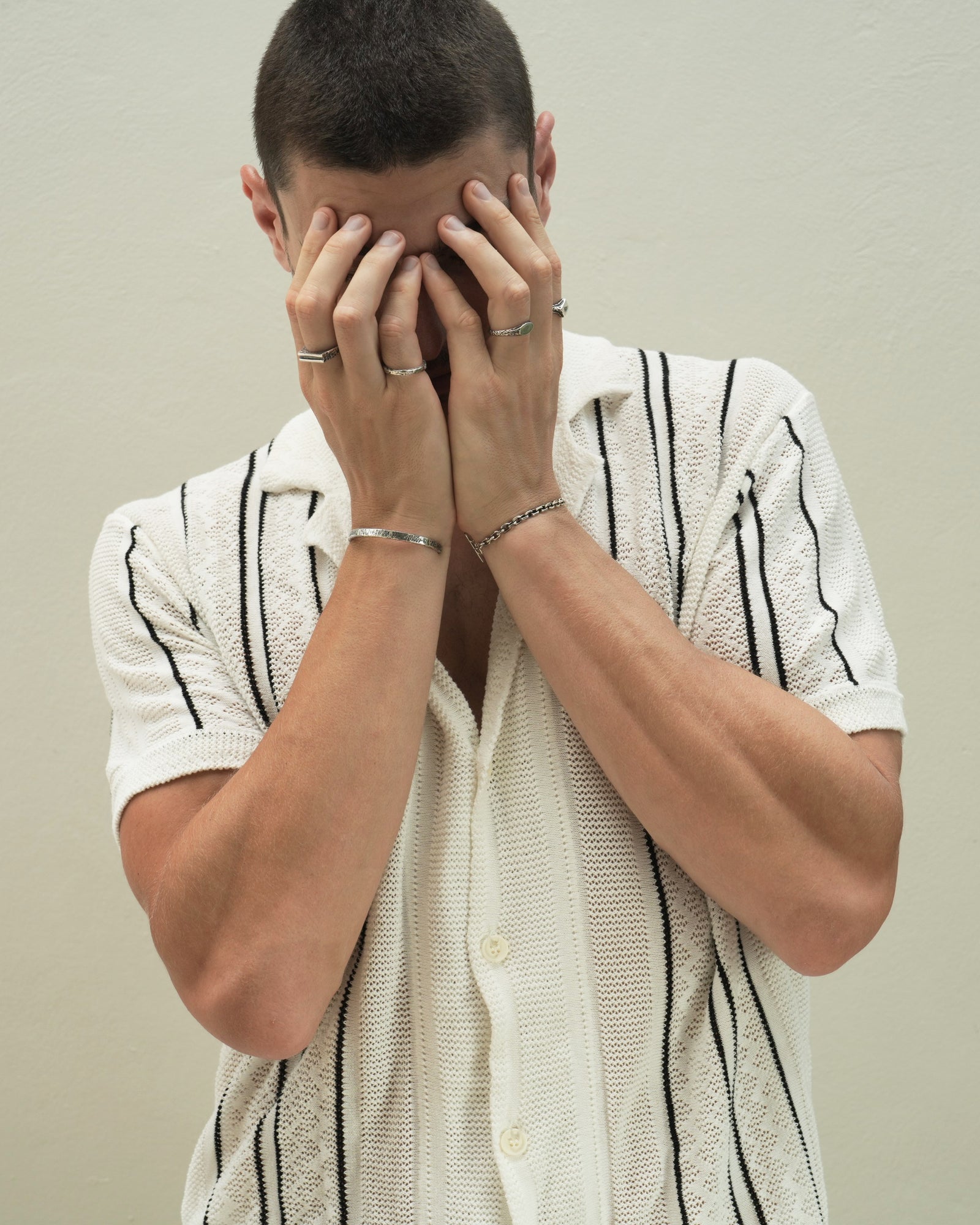 Founder of Nusands Corey Micari waist up front on view with both of his hands covering his face. Wearing sterling silver shackled bracelet on his left right wrist, mirrored signet and suns out rings on hand. Right arm wearing the striking cuf and line in the sands ring with stacker ring.