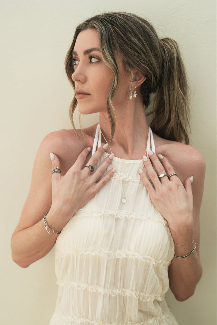 Founder of Nusands Emily Micari, looking away hands both up to chest wearing Sterling Silver earrings pearl and star charms in left ear, signature chain necklace with moon charm around neck, striking cuff and signature chain bracelet with mirrored signet and stacker ring on left arm, shackled bracelet, blade and suns out rings on right hand.