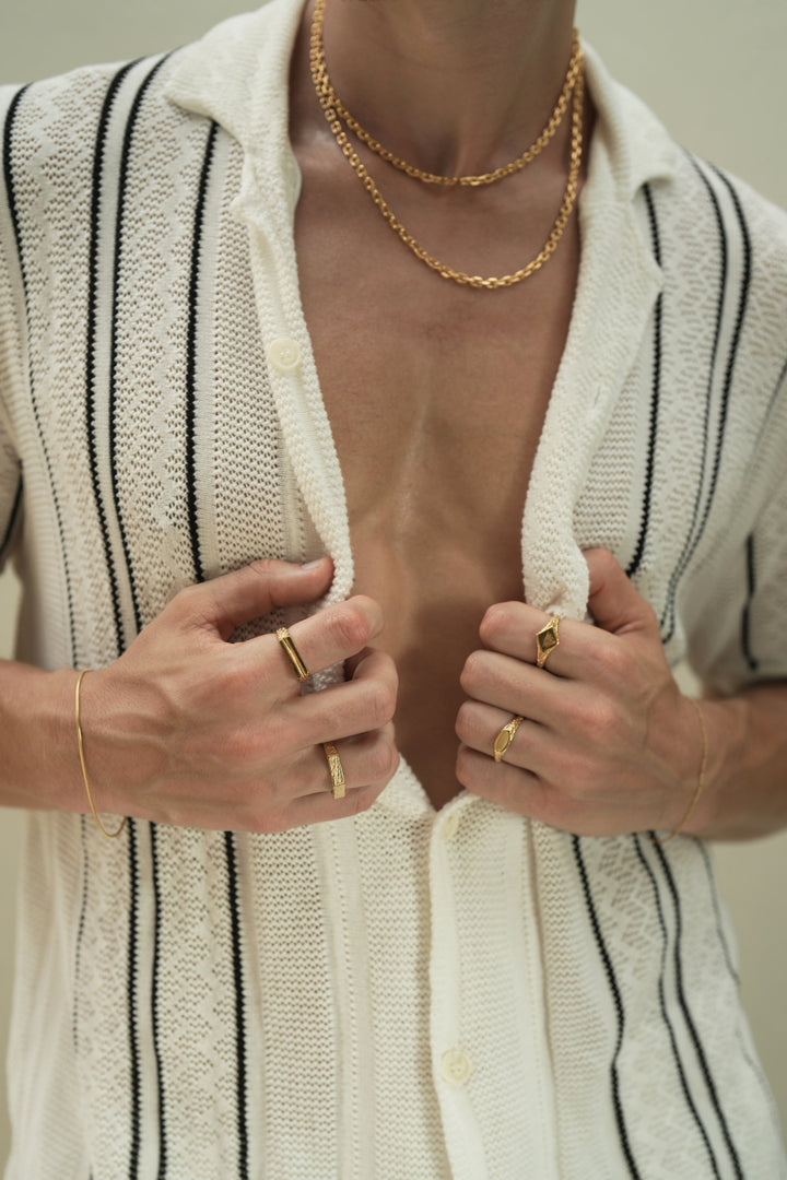 Founder of Nusands Corey Micari front view of chest wearing 18k gold layered shackled necklaces, hands both grasping the inside of button up shirt at chest hieght. Right hand wearing roped in bracelet, line in the sand and six base rings, left hand wearing signature chain, suns out and mirrored signet rings.