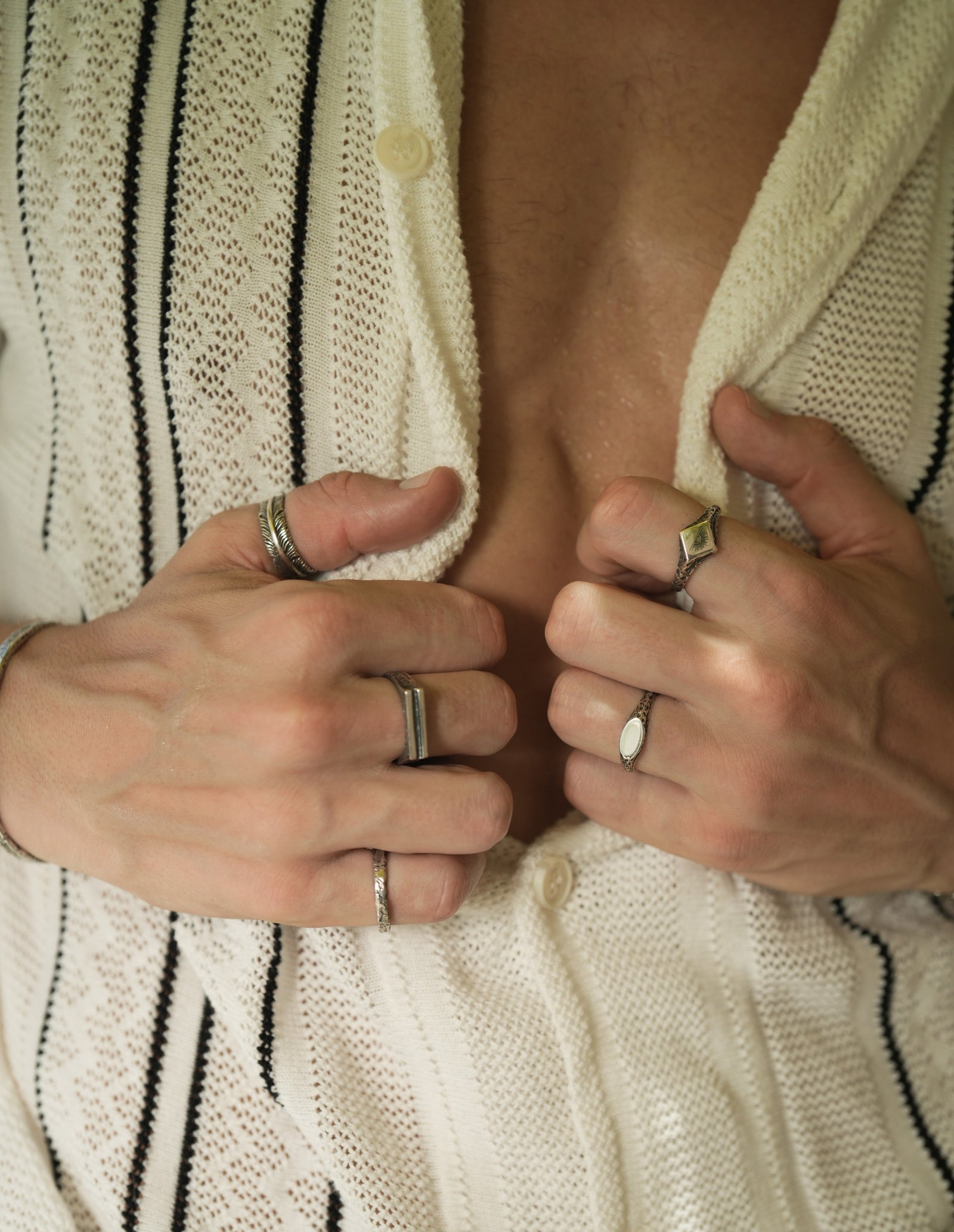 Founder of Nusands Corey Micari front view close up of chest, half unbuttoned white and black striped shirt both hands clasping the inside. Mirrored signet and suns out rings on left hand, striking cuff, blade, line in the sand and stacker rings on right hand.