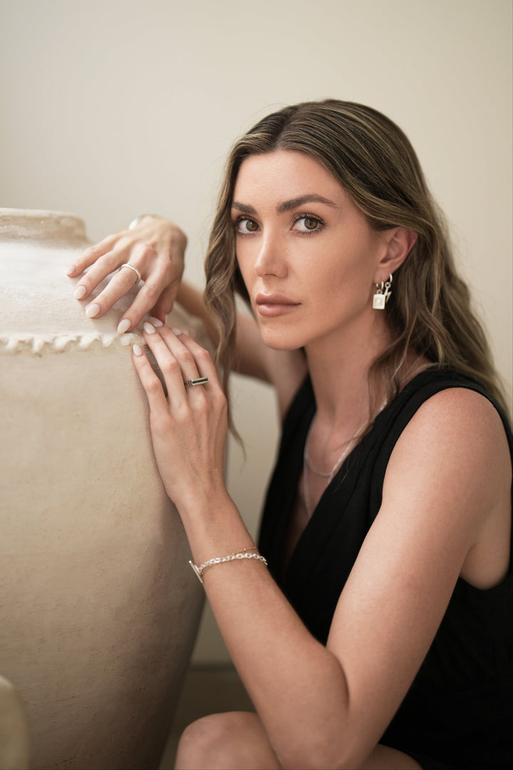 Founder of Nusands Emily Micari crouched next to a pot wearing sterling silver shackled bracelet and line in the sand ring on left hand, stacker ring on right, evil eye and lightning bolt earrings in left ear looking at camera.