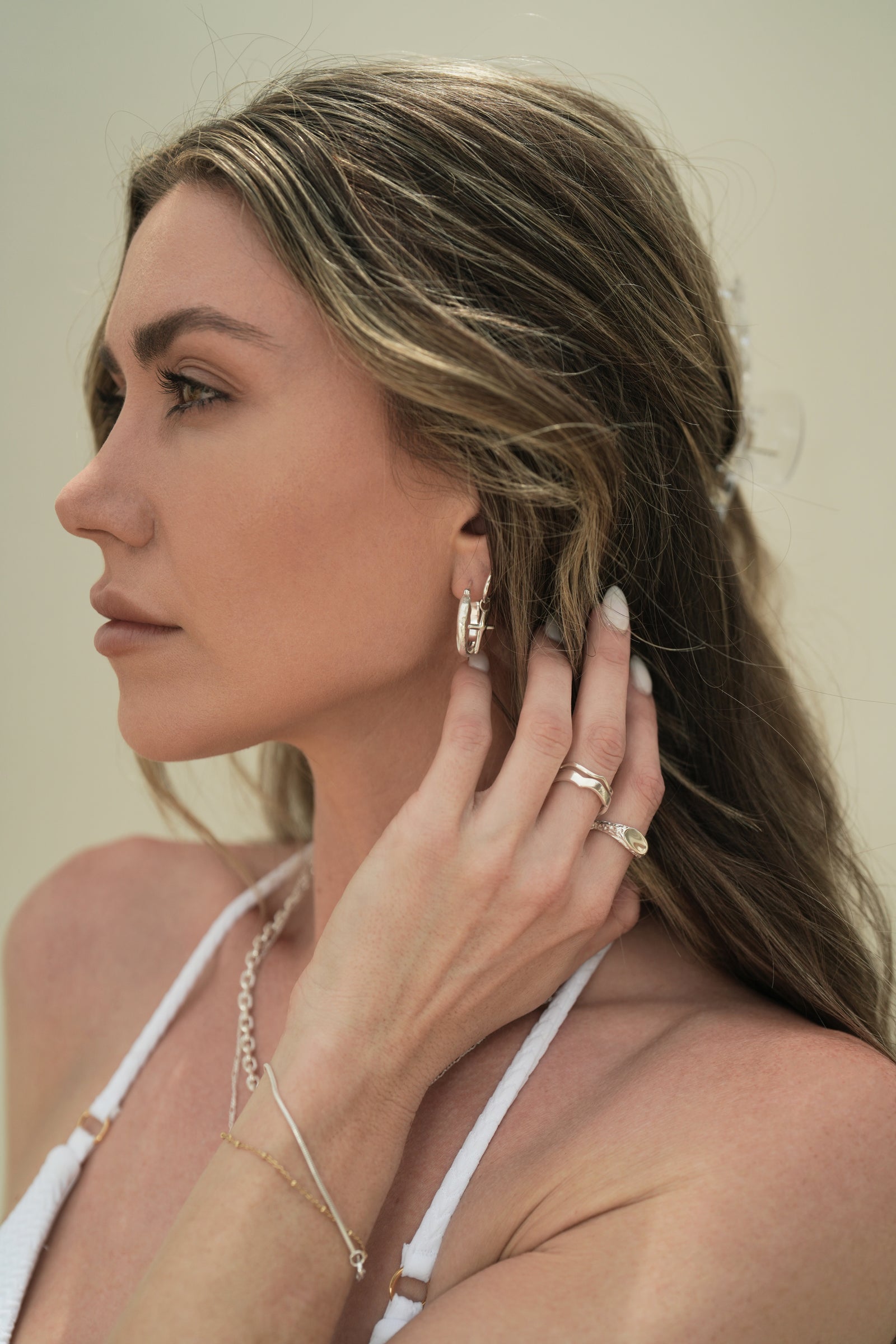 Founder of Nusands Emily Micari side profile of head wearing sterling silver stacked wavy and hoop earrings with cross charm in left eart. Left hand to side of neck showing stacked wave rings, mirrored signet ring, roped in bracelet and shackled necklace around her neck. 