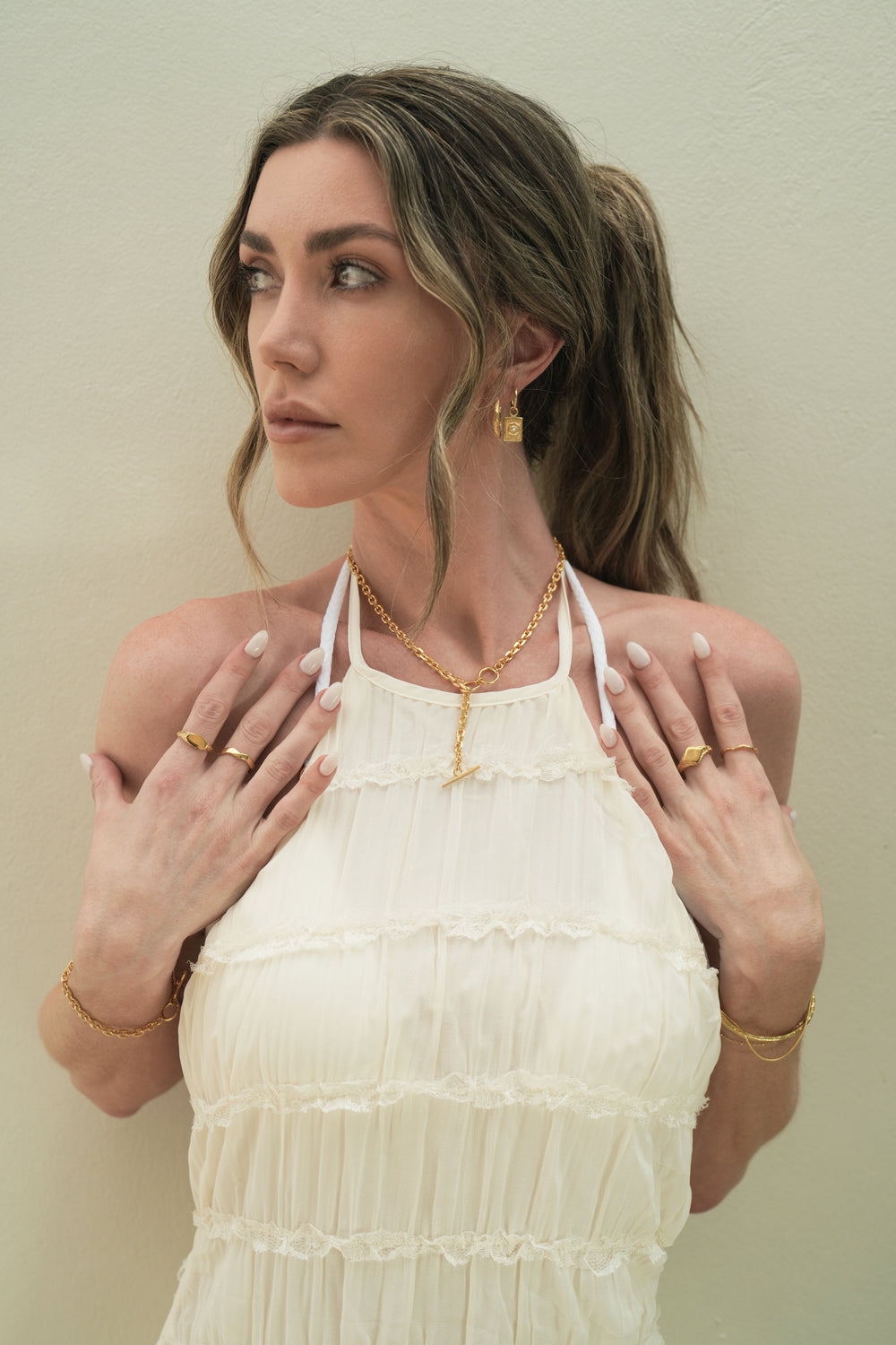 Founder of Nusands Emily Micari front on view hands on chest, looking away from camera. 18K gold Evil eye and wavy earrings stacked in left ear, striking cuff and signature chain with suns out and wave ring on left hand, shackled necklace on and shackled bracelet with mirrored signet and stacker ring on right hand.